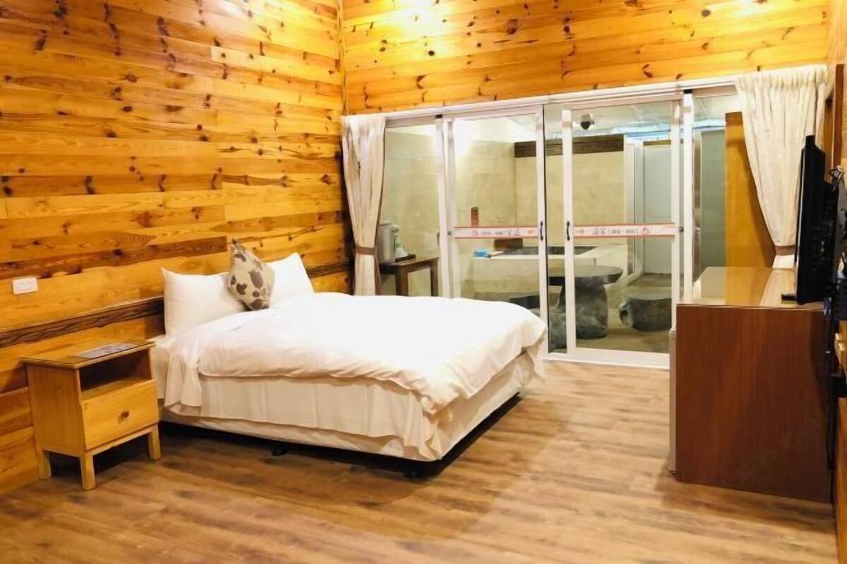 A wooden-paneled room with a bed, a small nightstand, a desk, and a sliding glass door leading to a bathroom.