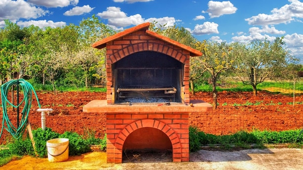 A brick-built outdoor oven with a sloped roof, surrounded by a garden with trees and a fence.