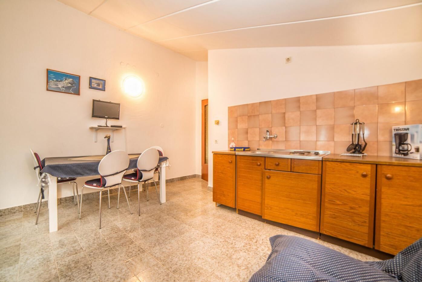Een kleine keuken met een eethoek, voorzien van een betegelde spatwand, houten kastjes en een tafeltje met stoelen.