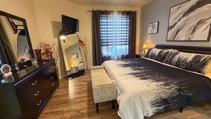 A bedroom with a bed, a dresser, a mirror, a TV, a window with curtains, and a wall with a painting.