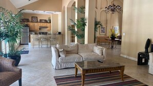 A spacious living area with a beige sofa, a coffee table, and a striped rug. There are potted plants and a chandelier.
