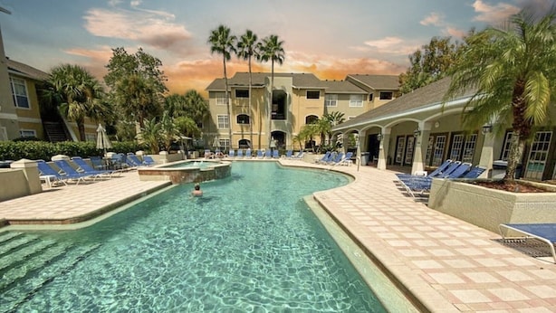 A swimming pool with clear water, surrounded by lounge chairs and palm trees, with a building in the background.