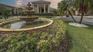 A building with a number '1220' on the facade, a fountain with multiple tiers, and a well-maintained garden with palm trees.