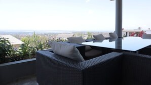 Een terras met rieten meubels, een tafel en uitzicht op een stadsgezicht.