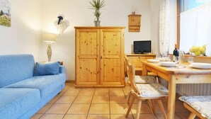 Una sala de estar con un sofá azul, armarios de madera, una mesa de comedor con sillas, un televisor y una ventana con cortinas.