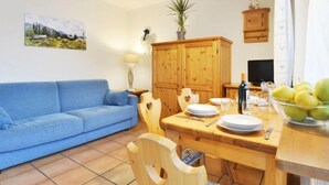 Una sala de estar con un sofá azul, una mesa de comedor de madera y sillas. Hay un cuadro en la pared, una lámpara y una planta sobre la mesa.