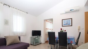 Un salón con un sofá morado, un televisor sobre un pedestal de cristal, una mesa de comedor con sillas y un cuadro en la pared.