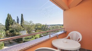 Un balcón con una mesa y sillas blancas, baranda de metal y vista a árboles y una carretera.