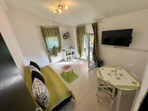 Ein kompakter Wohnbereich mit Sofa, Esstisch, an der Wand montiertem Fernseher und einer Glastür zum Außenbereich.