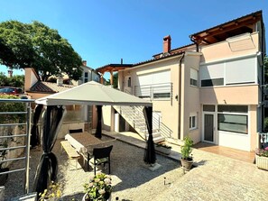 Ein Wohngebäude mit überdachter Terrasse, Außenbestuhlung und einer Treppe, die zu einer oberen Etage führt.