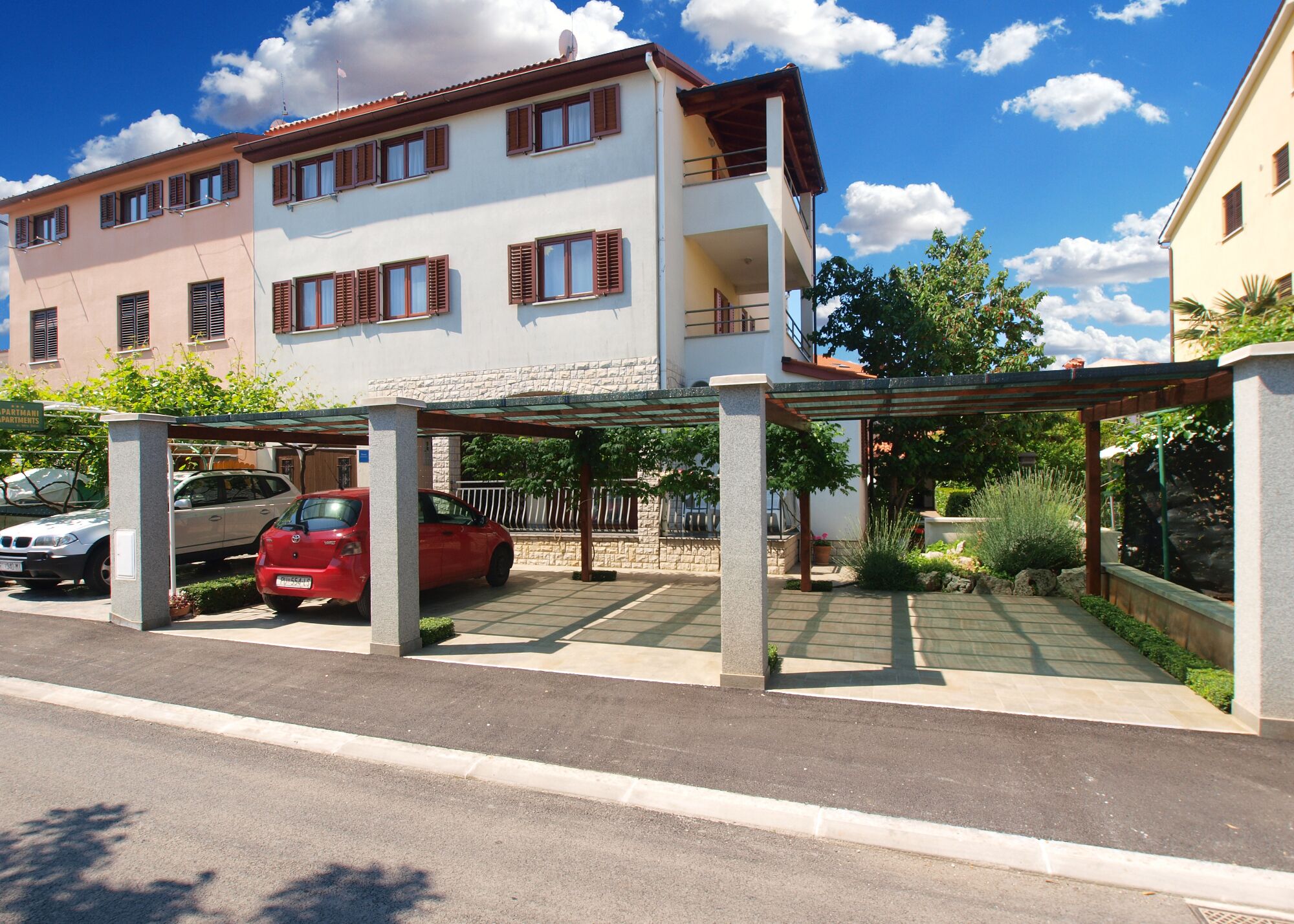 A multi-story residential building with a covered parking area, a red car parked inside, and a sign indicating 'PARTIMANI APARTMENTS'.