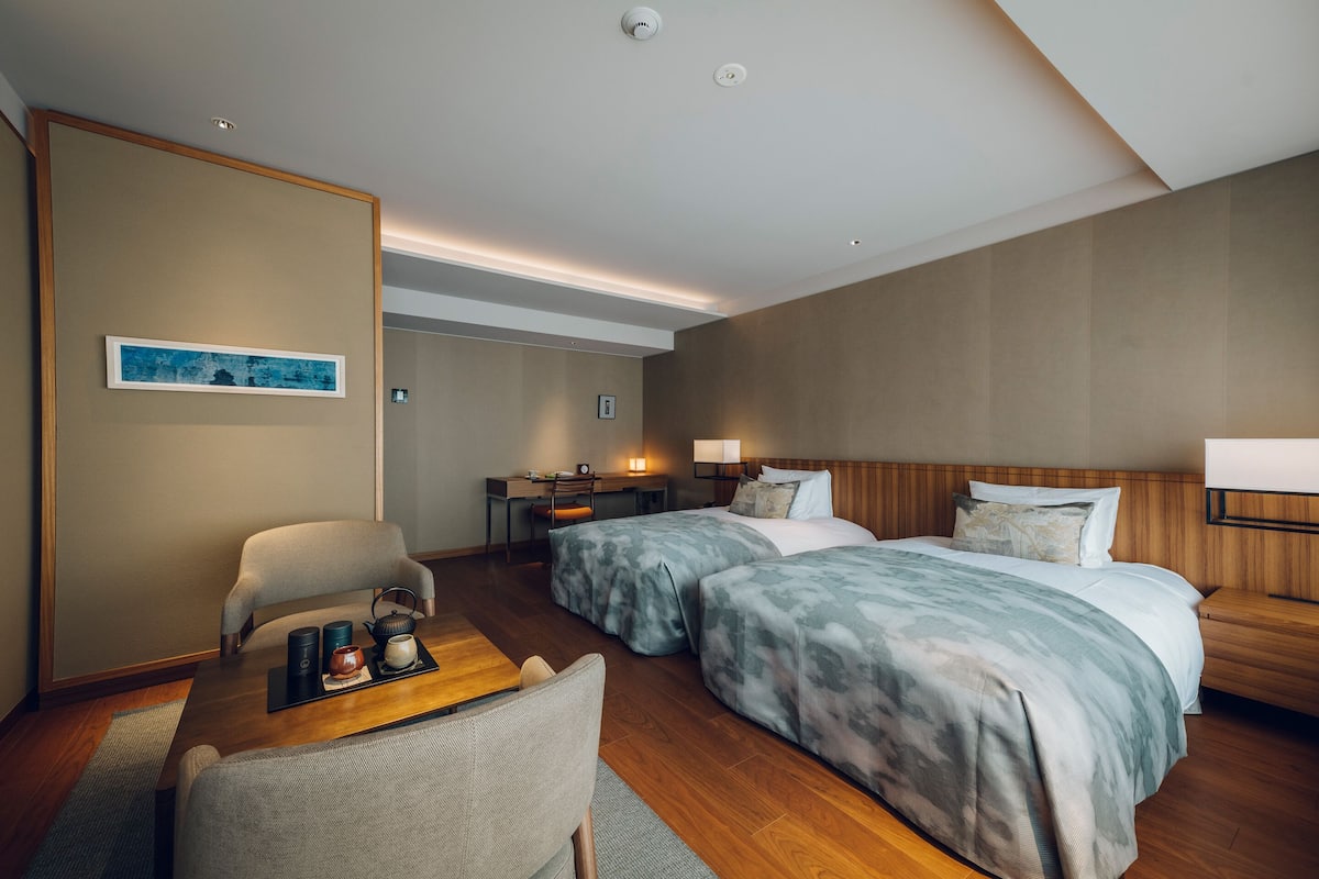 A hotel room with two beds, a desk, a chair, and a small table with a teapot and cups.