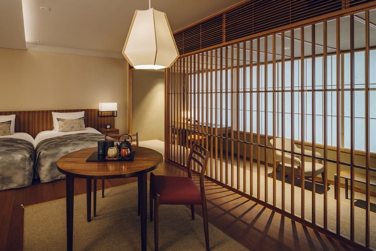A hotel room with a bed, a round table with a tea set, a chair, and a view through a wooden partition.