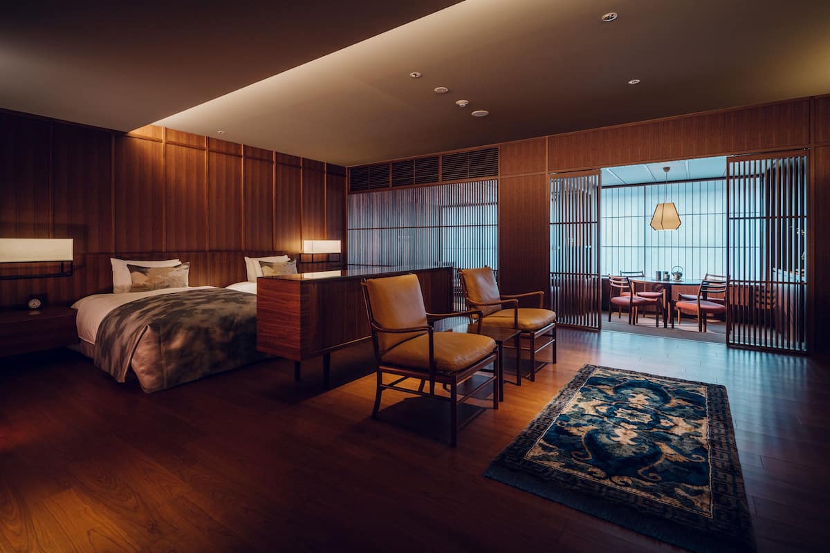 A modern hotel room with a wooden floor, a large bed, two chairs, a desk, and a view through a sliding door.