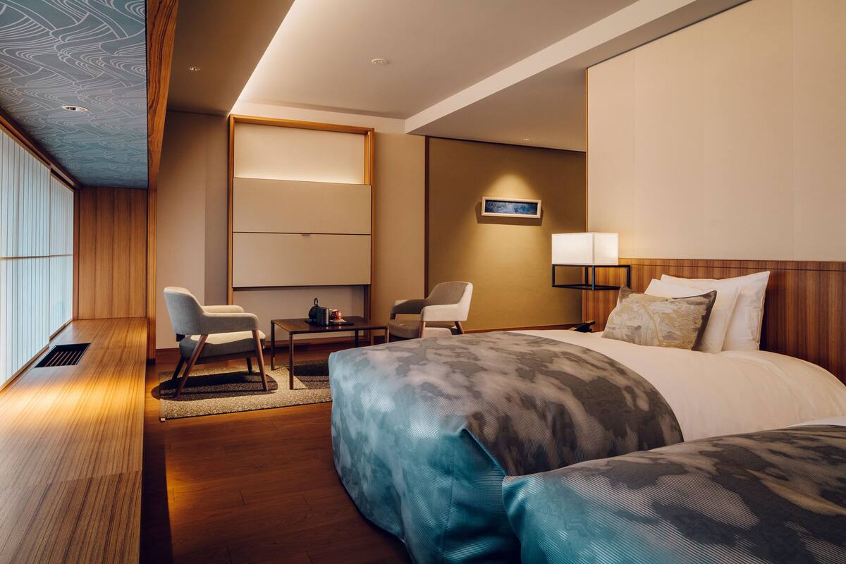 A modern hotel room with a large bed, a desk, two chairs, a TV, and a wall-mounted lamp.