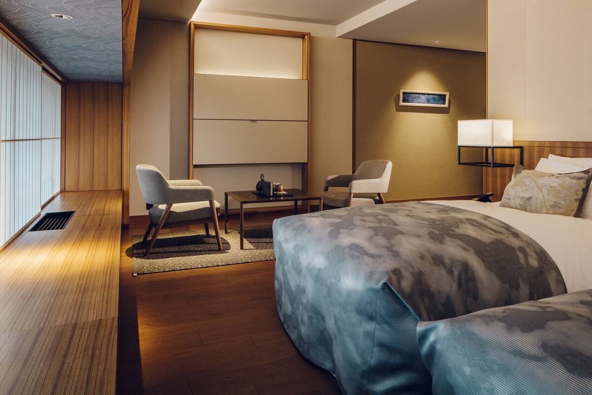 A hotel room with a bed, two chairs, a desk, a wall-mounted TV, and a bedside table.