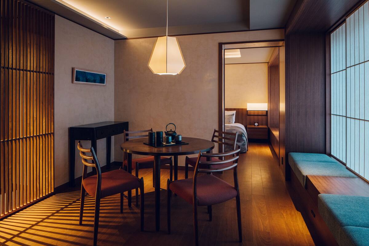 A traditional Japanese-style room with a tatami mat floor, a low table with chairs, a hanging light fixture, and a sliding door leading to a sleeping area with a futon bed.