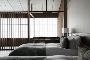 A modern hotel room with a large bed, a desk, and a view through a window with vertical blinds.