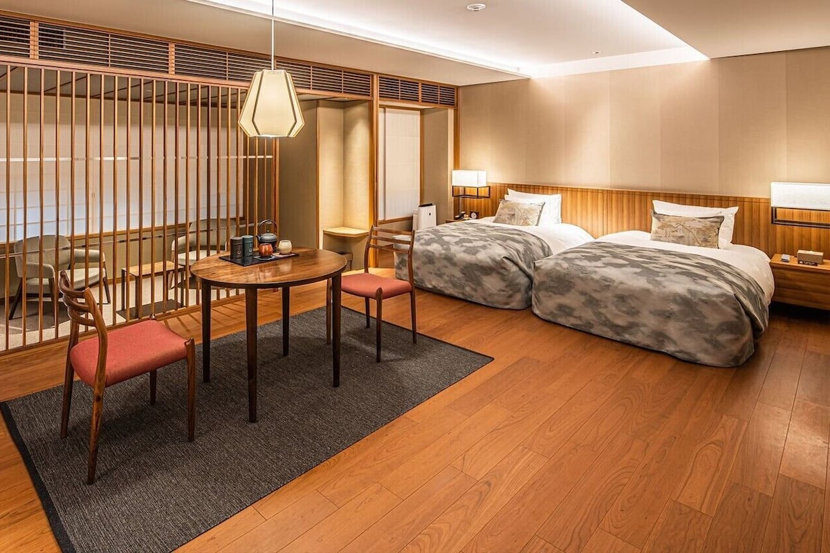 A hotel room with a wooden floor, a bed with a headboard, a desk with a chair, a table with chairs, and a partition with vertical slats.