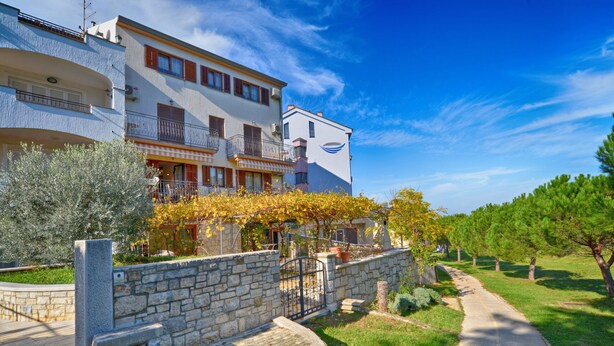 A multi-story hotel with balconies, a stone wall, and a pathway leading to a grassy area with trees.