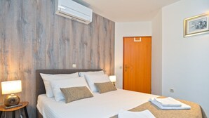 A neatly made bed with white linens and two pillows, a wooden wall panel, an air conditioning unit, a wooden door, and a framed picture on the wall.