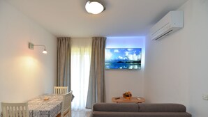 A living room with a sofa, a dining table with chairs, a TV on the wall, and an air conditioning unit.