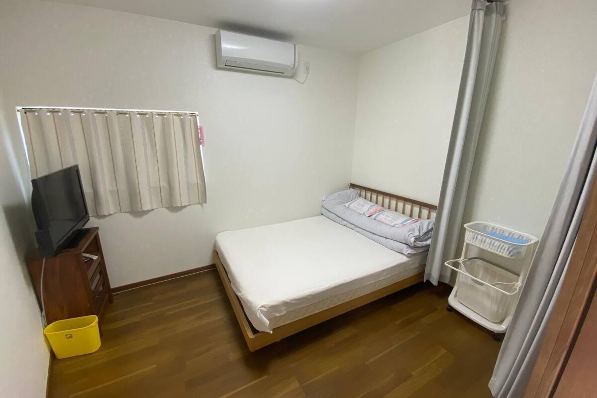 Ein kleines, sauberes Zimmer mit einem Bett, einem Fernseher auf einem Holzboden, einer Klimaanlage und einem Wäschekorb.