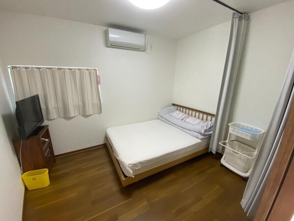 Een kleine, schone kamer met een bed, een televisie op een houten standaard, een airconditioner en een wasmand.