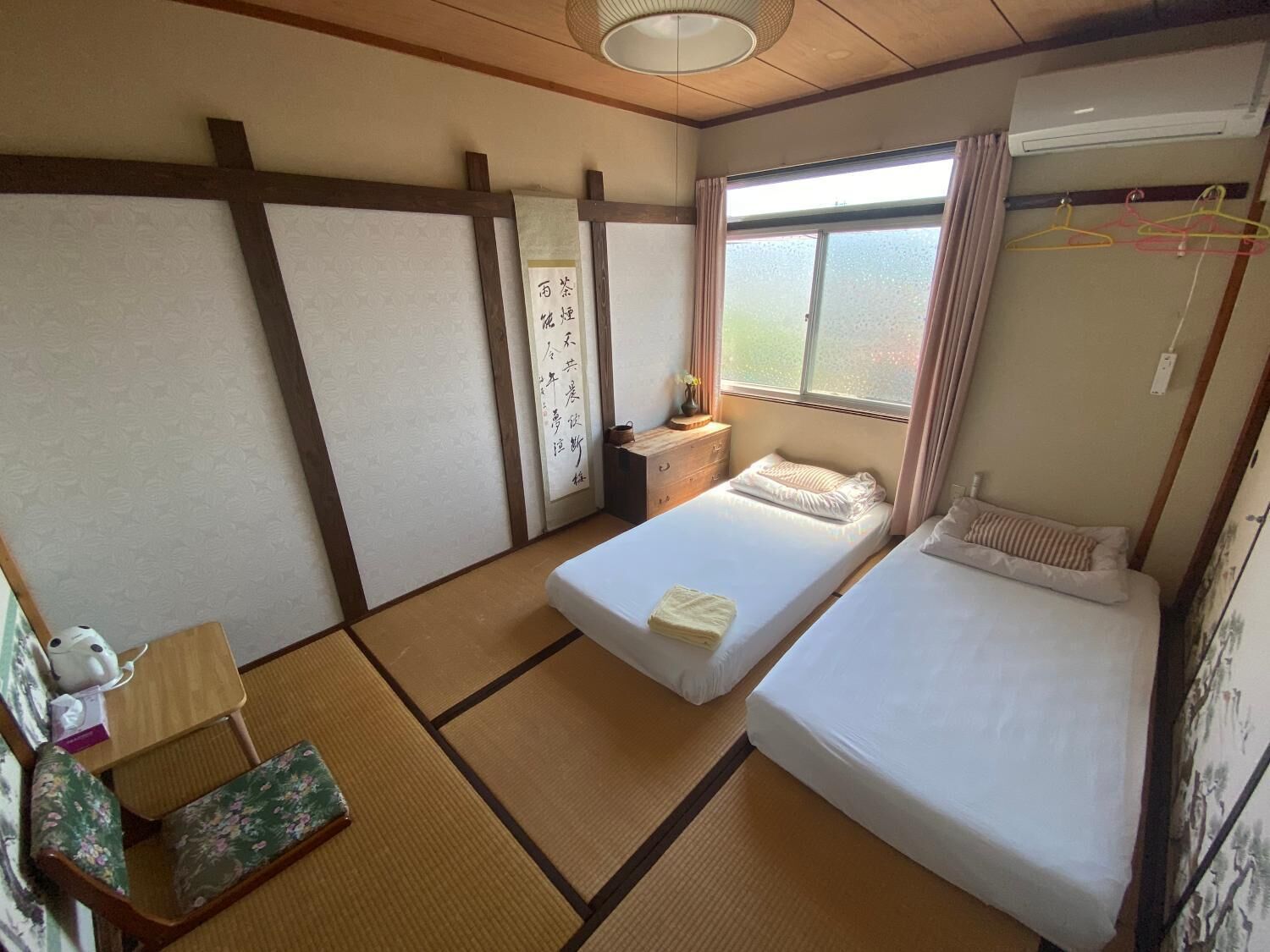 A traditional Japanese-style room with tatami flooring, two beds, a small table, and a window with a view.