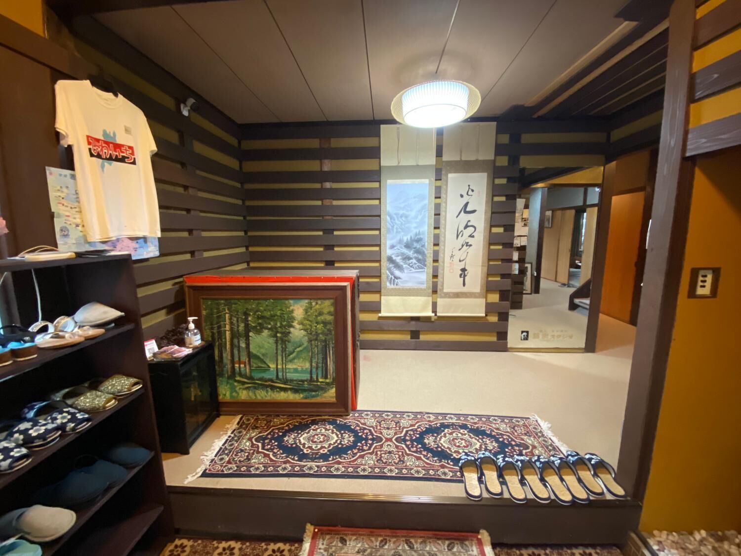 A room with a wooden slat wall, a framed painting of a forest, a patterned rug, and a shelf with shoes.