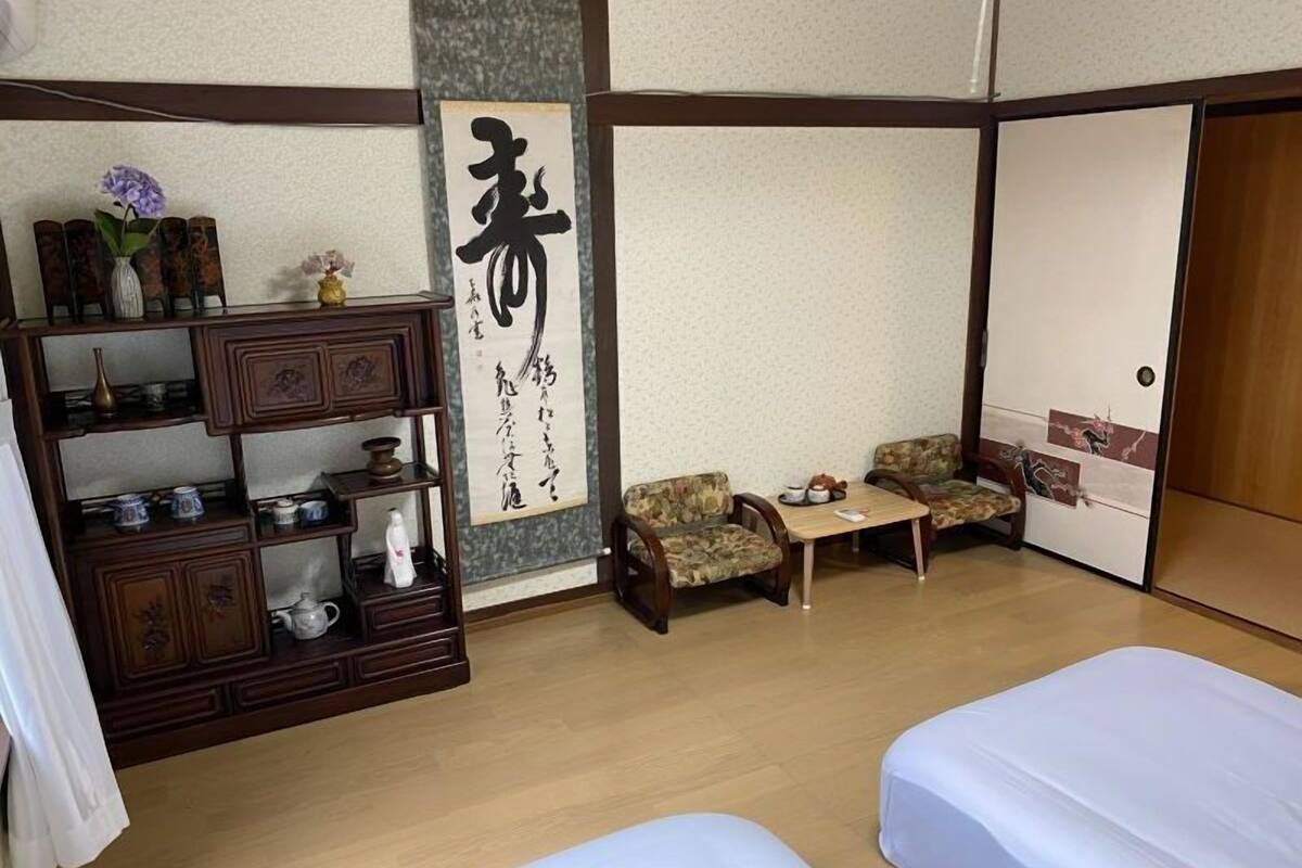 Ein traditioneller japanischer Raum mit Tatami-Bodenbelag, einem niedrigen Holztisch, zwei Tatami-Matten als Sitzgelegenheit, einem Regal mit Dekoration und einer Schiebetür.