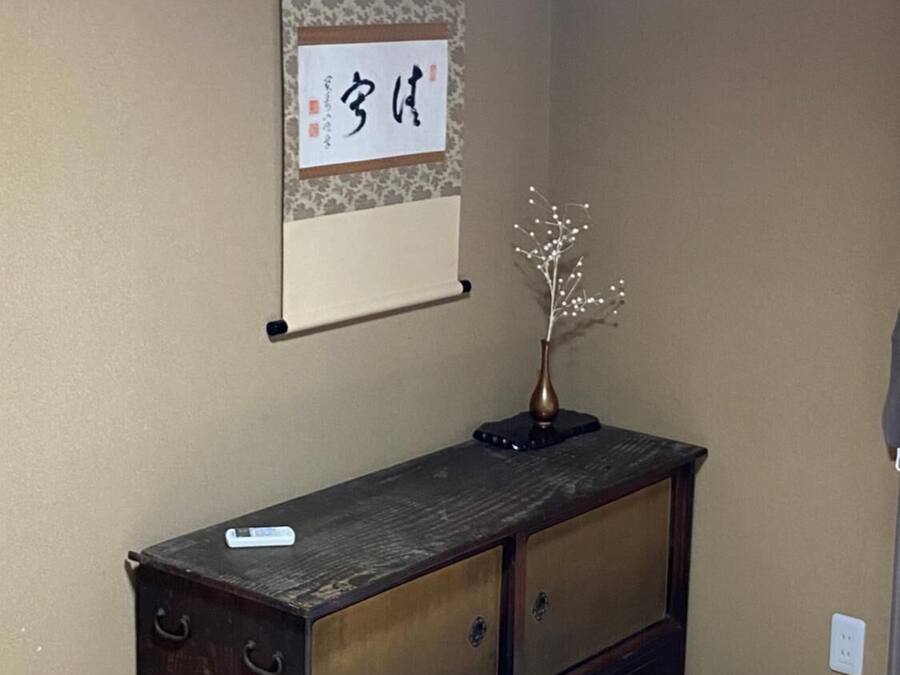 A bedroom with a bed, a dresser, a wall-mounted shelf with a decorative item, and a wall with a hanging sign.