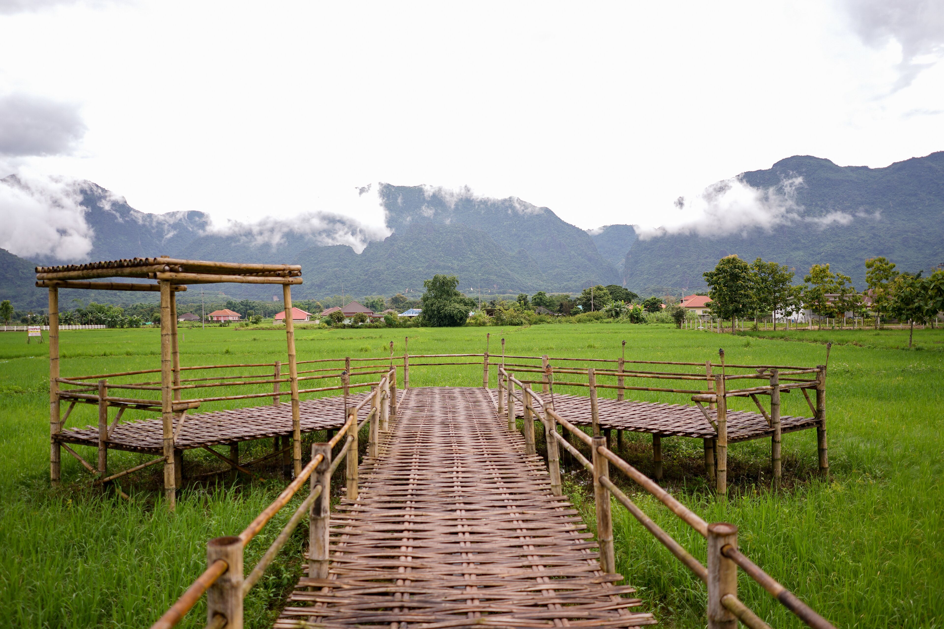 A rustic wooden bridge leading to a bamboo pavilion in a lush green field with mountains in the background.