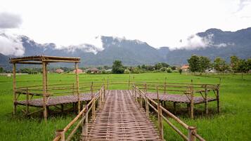 A rustic wooden bridge leading to a bamboo pavilion in a lush green field with mountains in the background.