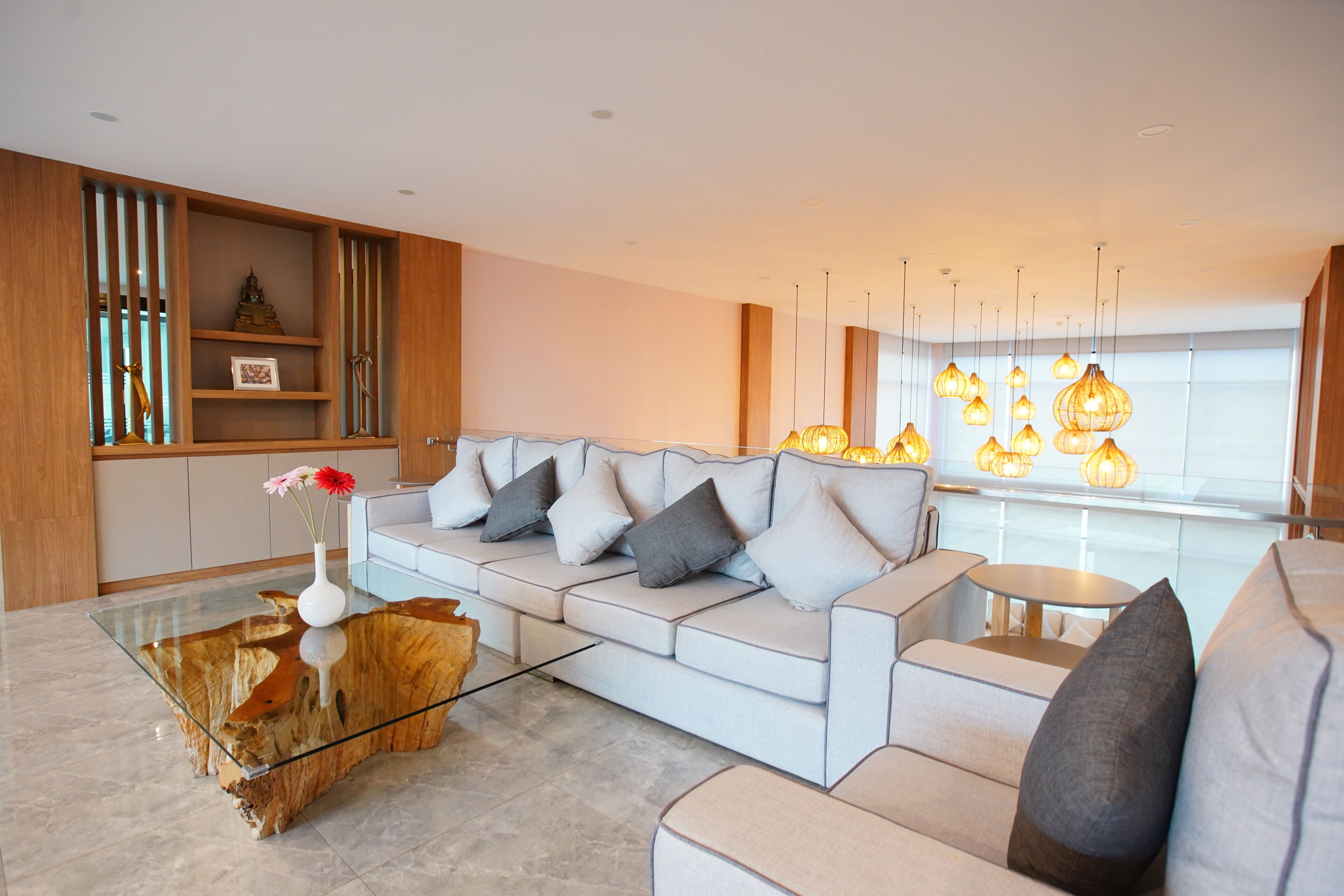 A modern living room with a glass coffee table, a grey sofa with cushions, wooden shelving, and pendant lighting.