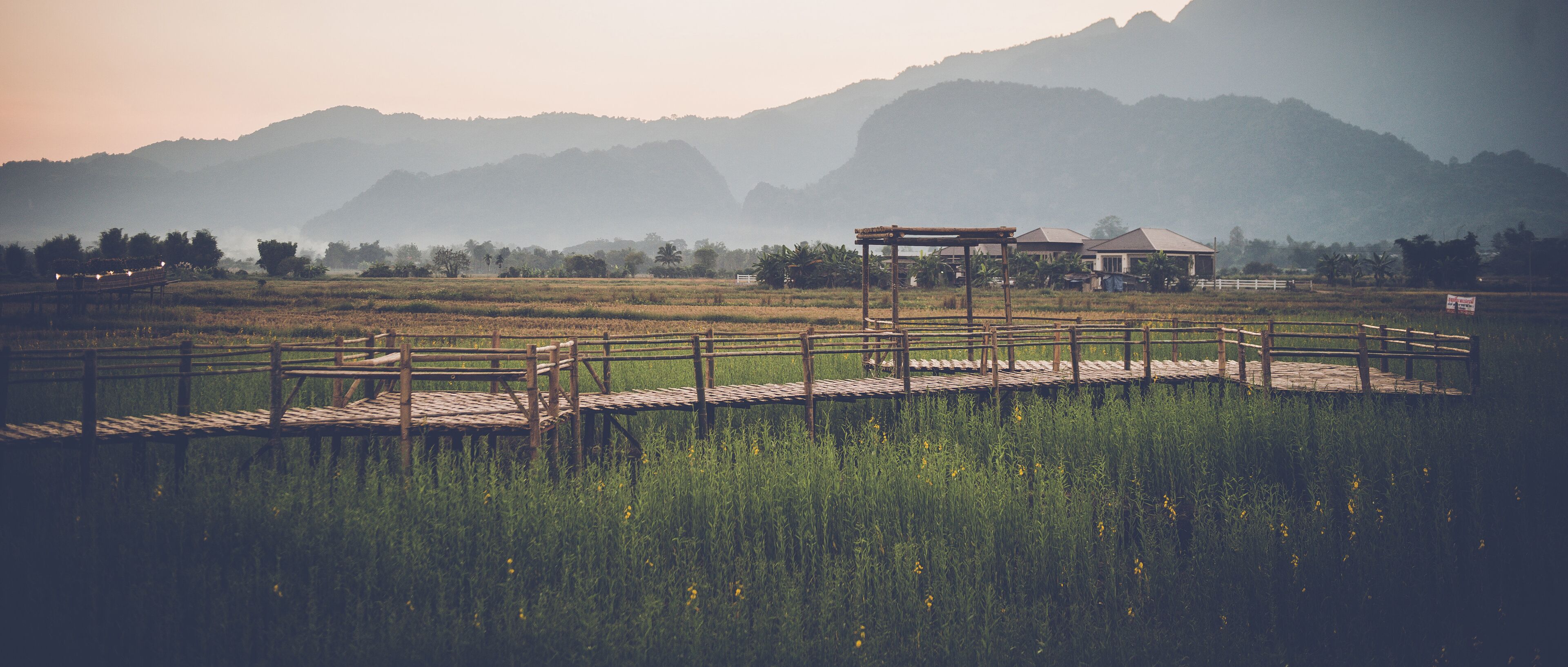 Een houten brug in landelijke stijl die leidt naar een tuinhuisje in een weelderig veld, met bergen op de achtergrond.