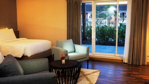Een hotelkamer met een bed, een bank, een stoel, een klein tafeltje met bloemen en uitzicht op een zwembad en groen door een groot raam.