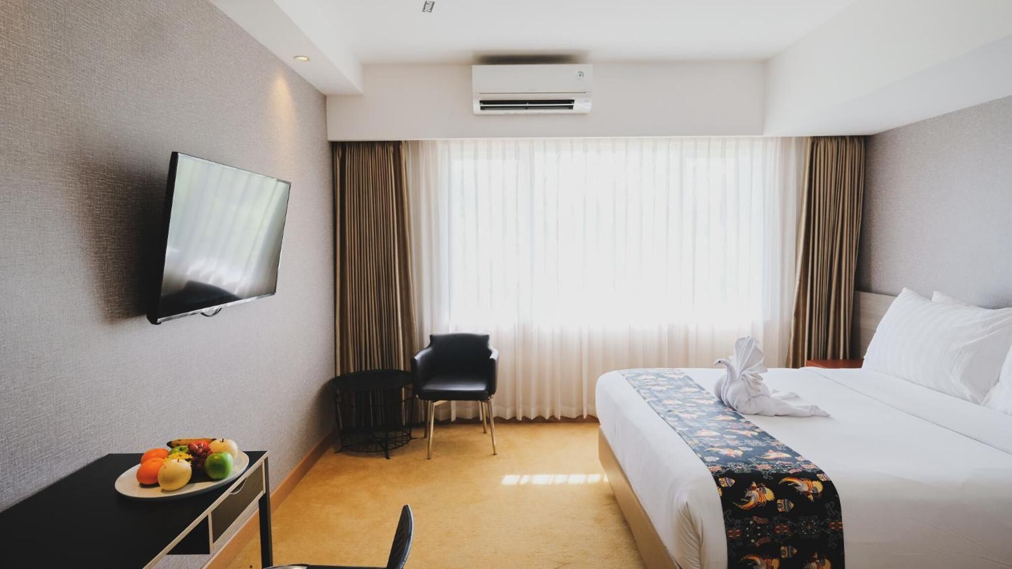 A hotel room with a bed, a TV mounted on the wall, a desk with fruit, a chair, and a window with curtains.