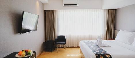 A hotel room with a bed, a TV mounted on the wall, a desk with fruit, a chair, and a window with curtains.