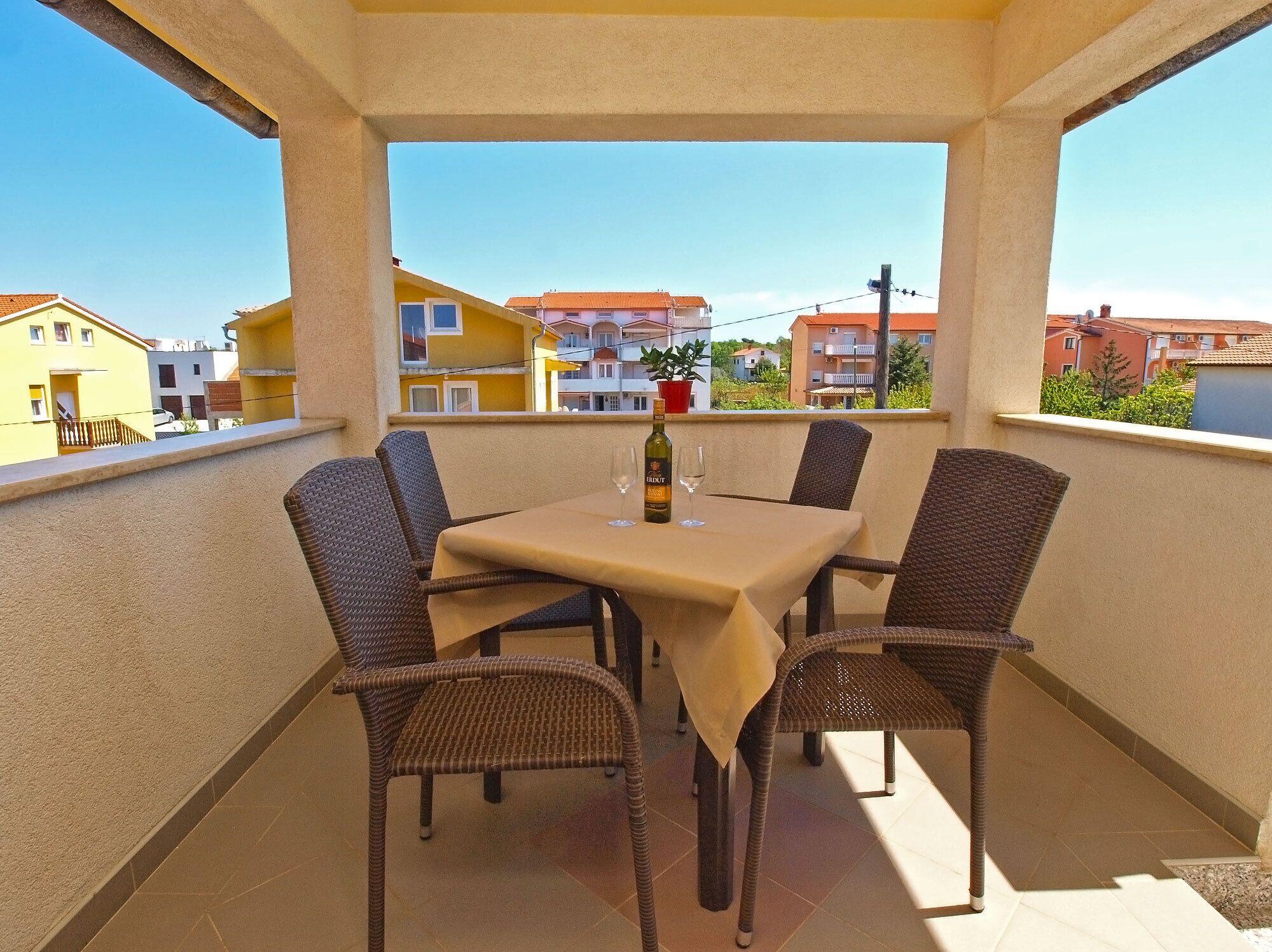 A balcony with a table and four wicker chairs, overlooking a residential area with colorful buildings.