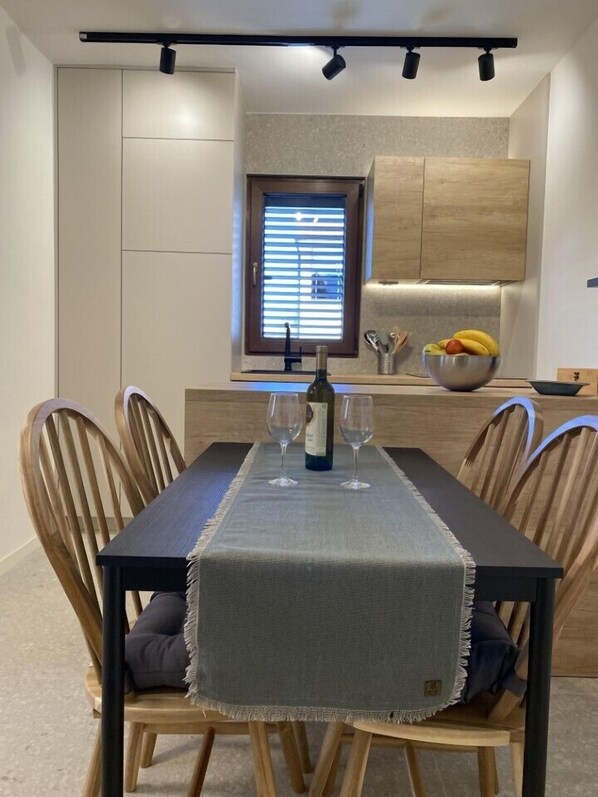 A dining area with a table set for two, a bottle of wine, and two glasses. There are wooden chairs with cushions, a window with blinds, and a kitchen area with cabinets and a fruit bowl.