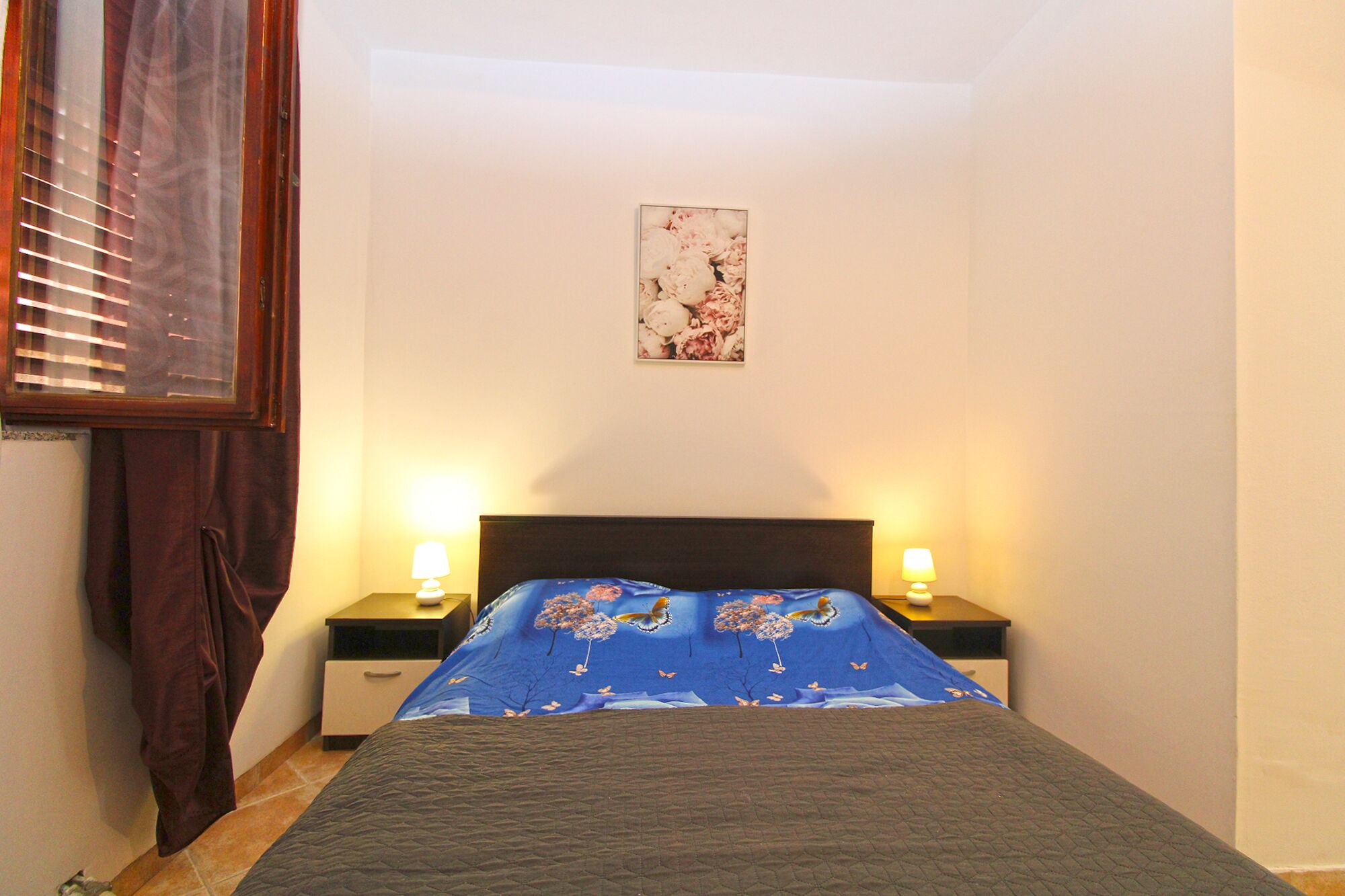 A bedroom with a bed, two bedside lamps, a nightstand, a window with blinds, and a wall-mounted picture.