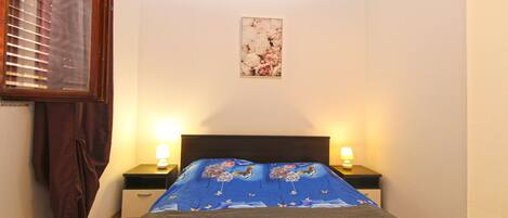 A bedroom with a bed, two bedside lamps, a nightstand, a window with blinds, and a wall-mounted picture.