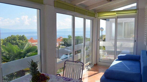 A balcony with a table and chairs, a blue sofa, and a view of the sea and greenery.