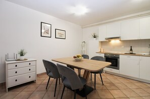 A modern kitchen with white cabinets, a wooden dining table, and two chairs. There is a bowl of fruit on the table.
