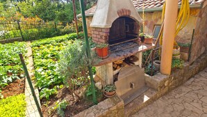 An outdoor brick oven with a stone chimney, surrounded by a garden with various plants and a fence. - Apartment 1348/12670 (Istria - Pula), 100m from the beach (Pula)