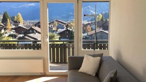 A modern living room with a grey sofa, a white furry throw, and a view of a mountainous village through large windows. - Turmalin 2. Stock by Interhome (Lenk)