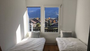 A small room with two beds, a wooden floor, and a balcony with a view of a mountain and buildings. - Turmalin 2. Stock by Interhome (Lenk)