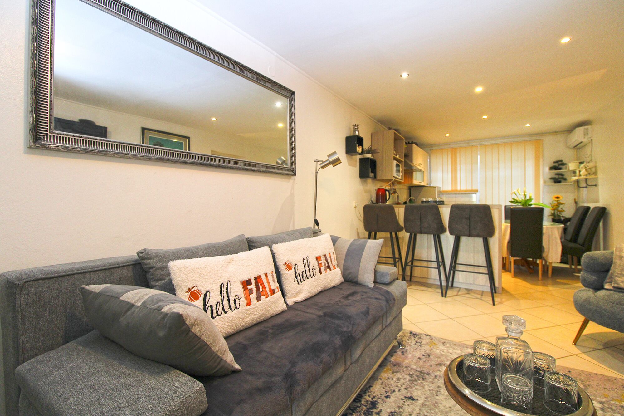 Una sala de estar con un sofá gris, una mesa baja con bebidas, una cocina al fondo y un espejo grande en la pared.