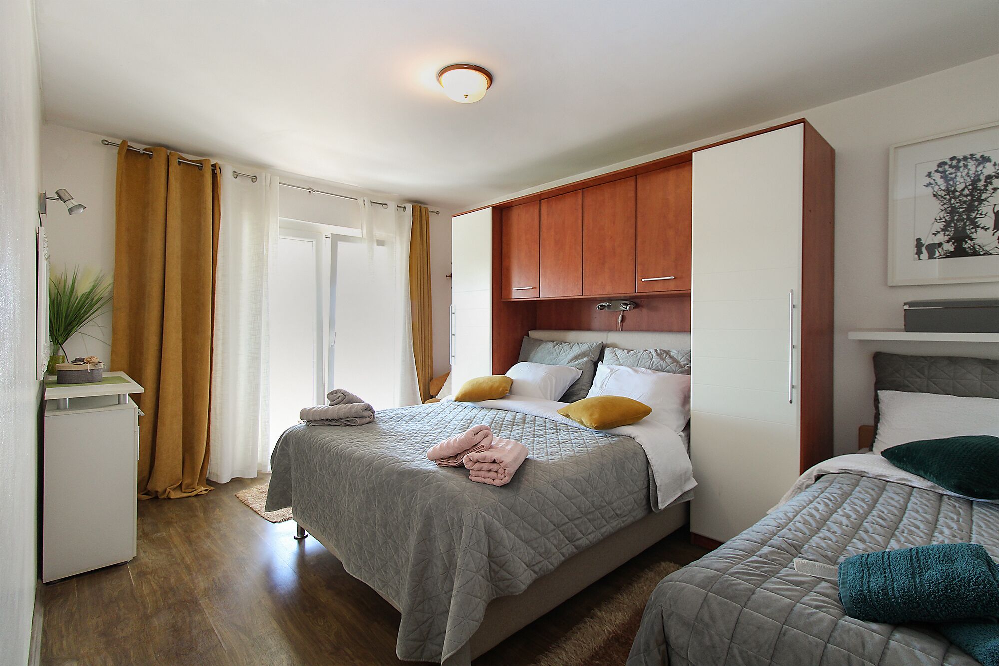 A bedroom with a bed, a smaller bed, wooden cabinets, a desk, and a plant.